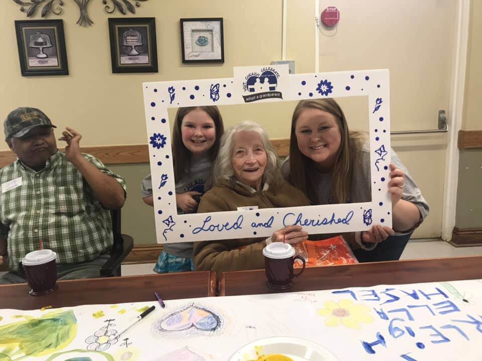Three generations smiling: an elderly woman, a young girl, and a young woman holding a 'Loved and Cherished' frame at an 'Adopt a Grandparent' event.