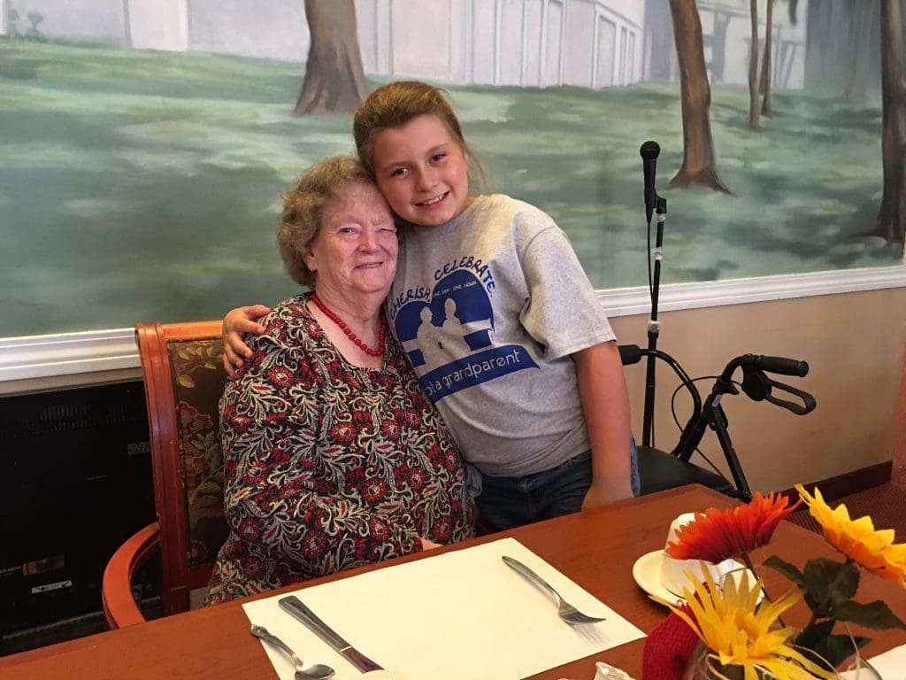 Smiling grandmother and granddaughter hugging at a dining table. The girl's shirt, 'Cherish Celebrate Be a Grandparent', beautifully depicts their loving family connection.